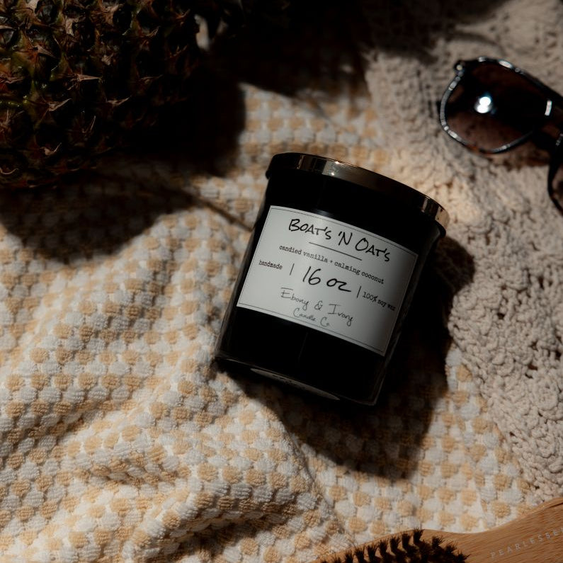 A black 16oz soy candle with a gold lid, labelled Boats N Oats, laying on a beach blanket with sunglasses and a pineapple. Made by Ebony & Ivory Candle Co in Lethbridge Alberta Canada