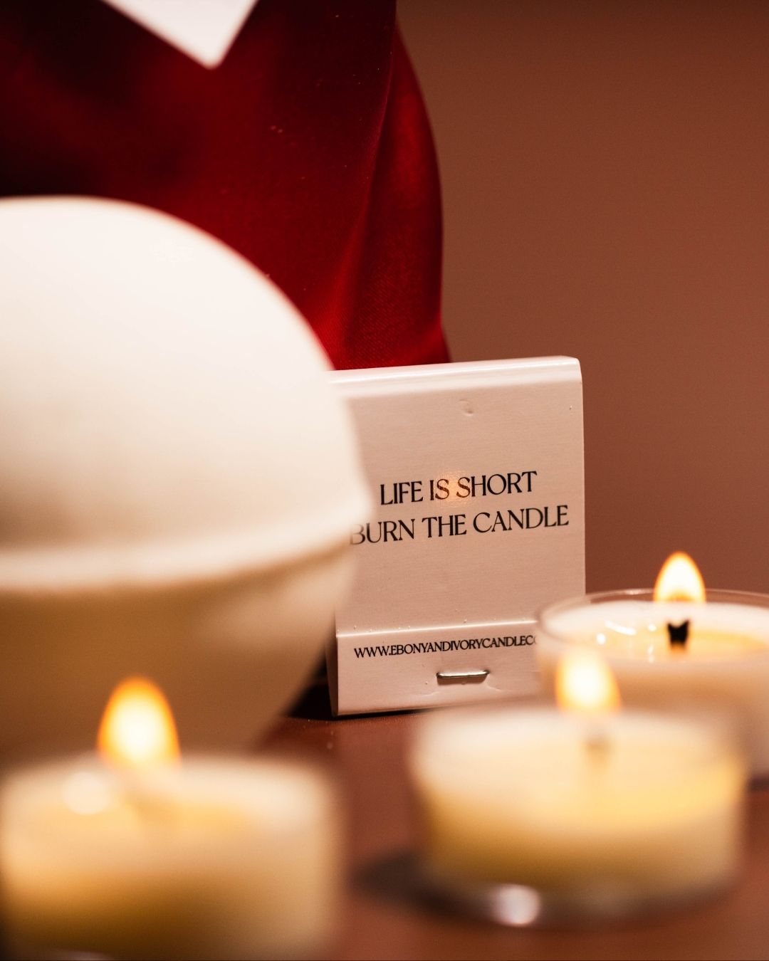 A book of matching sitting with a white christmas bath bomb infront of a red satin bag and tea lights.