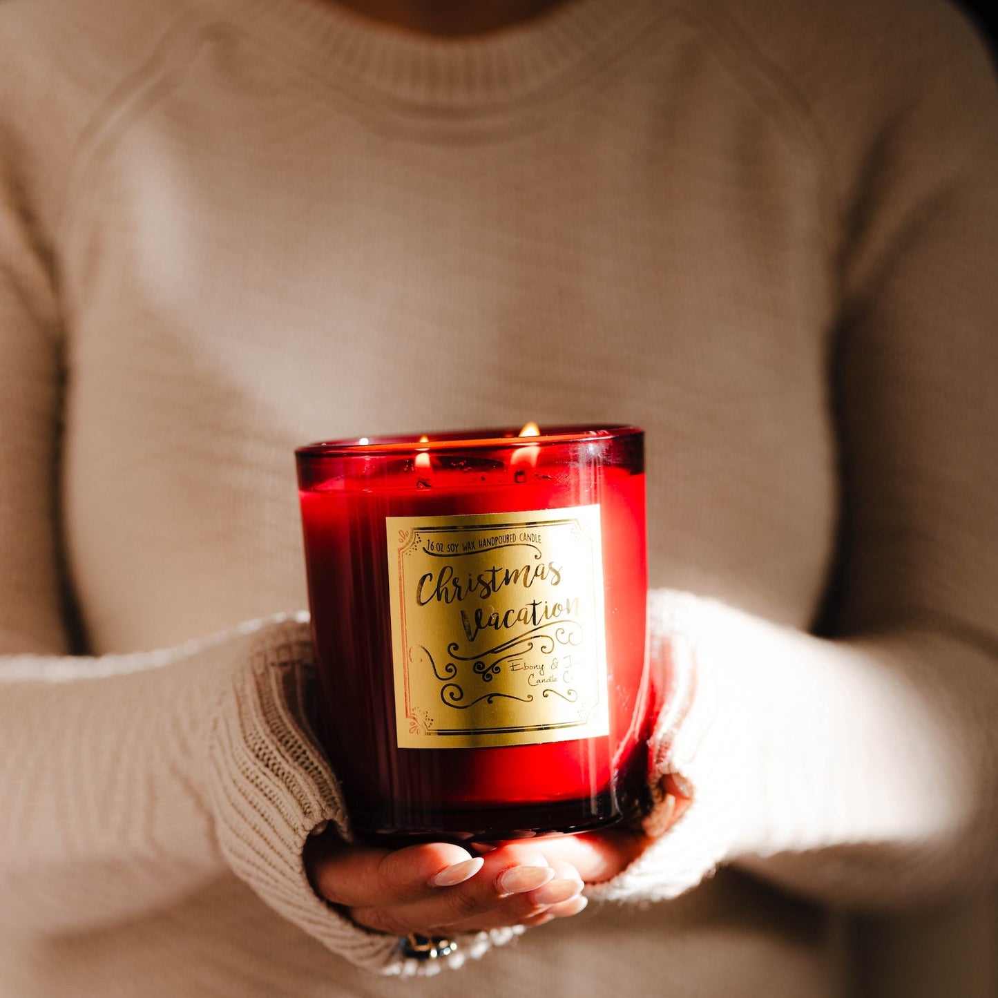 Person holding a red jar Christmas soy candle with a gold label, wearing a beige sweater. Eggnog scented candle called Christmas Vacation.