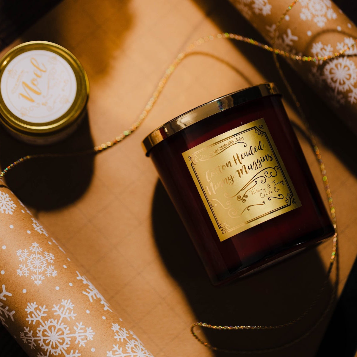Christmas soy Candle with a decorative label on a surface with gift wrap. Snowflake wrapping paper with velvet green ribbon. Best Christmas candle Canada. Gift for her. Cotton Headed Ninny Muggins candle.