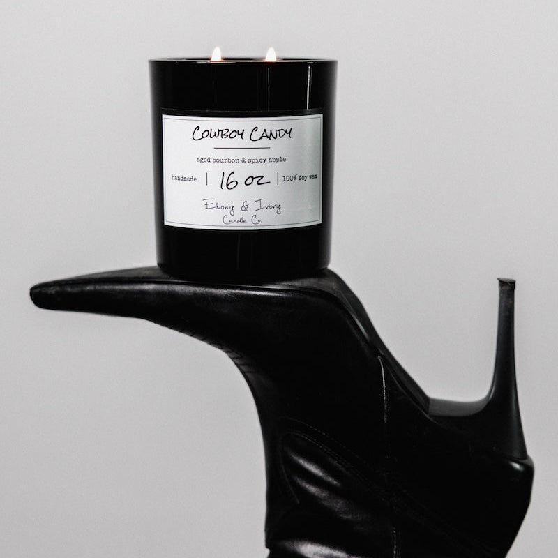 A black 16oz jar soy candle with a white label named Cowboy Candy on a black high heeled boot on a background, made by Ebony & Ivory Candle Co in Lethbridge, Alberta, Canada