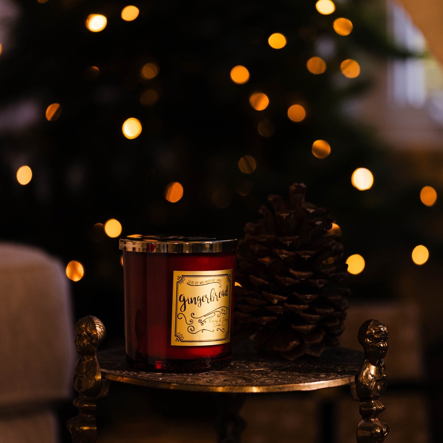 Red candle with a label on a small table in front of a decorated Christmas tree with lights. Gingerbread soy wax Christmas candle with red jar and gold label.