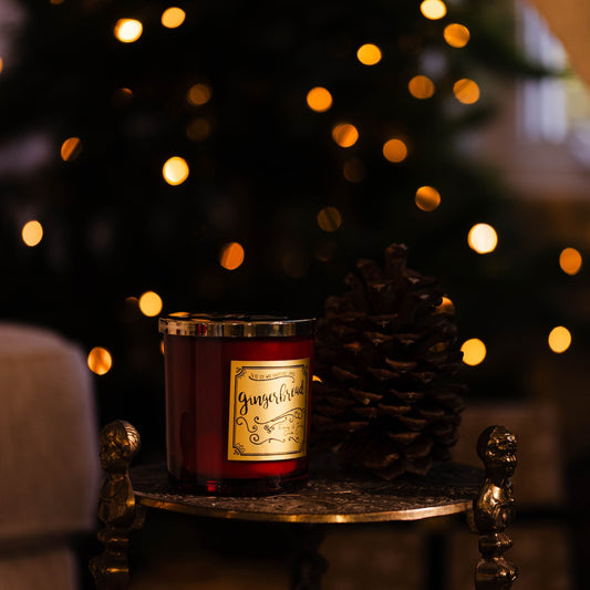 Red candle with a label on a small table in front of a decorated Christmas tree with lights. Gingerbread soy wax Christmas candle with red jar and gold label.