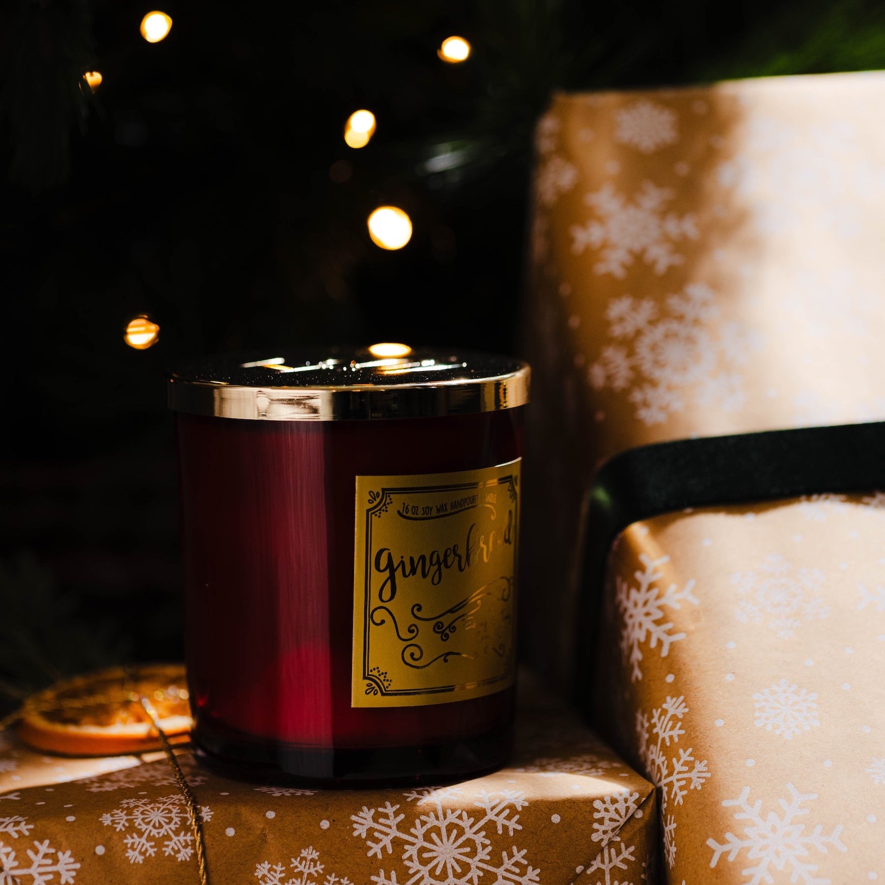 Ginger candle on a stack of wrapped presents with a Christmas tree in the background. Red candle with a label on a small table in front of a decorated Christmas tree with lights. Gingerbread soy wax Christmas candle with red jar and gold label.