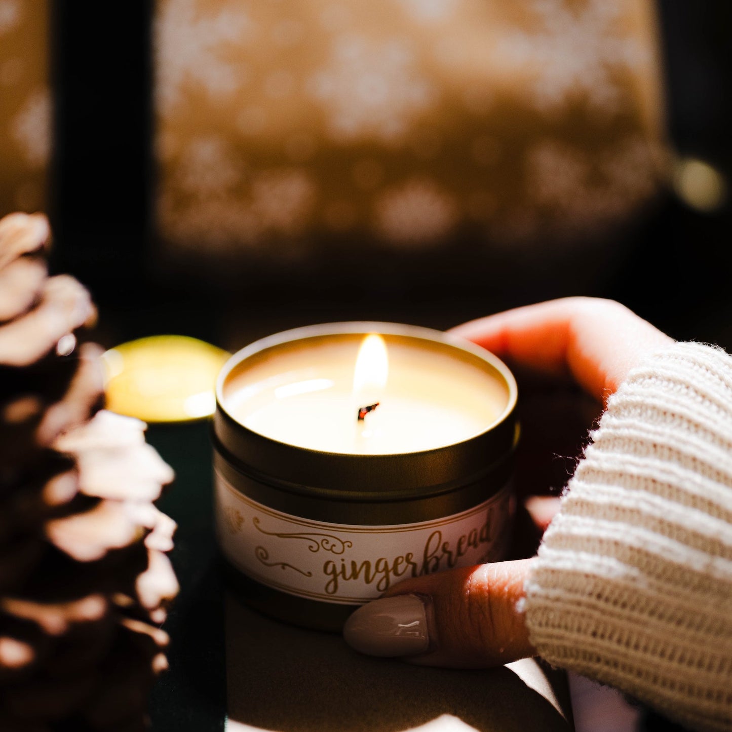 Gold tin candle with a label on a small gift in front of a decorated Christmas tree with lights. Gingerbread soy wax Christmas candle.