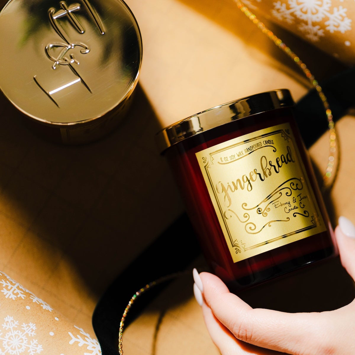 Red candle with a label on a small table in front of a decorated Christmas tree with lights. Gingerbread soy wax Christmas candle with red jar and gold label. Christmas snowflake wrapping paper with green velvet ribbon and gold twine.
