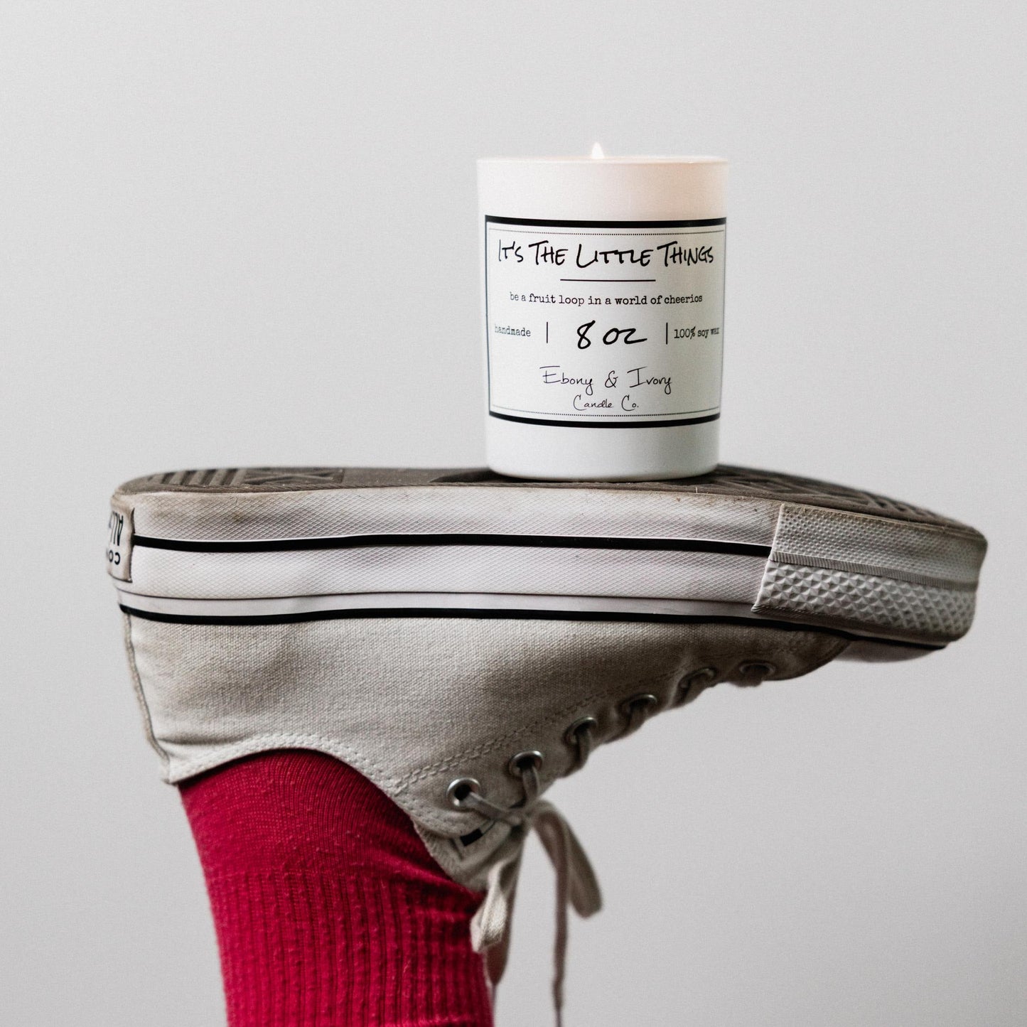 A white jar 8oz soy candle with a gold lid and a white label named It's The Little Things on a white sneaker with a red sock, made by Ebony & Ivory Candle Co in Lethbridge, Alberta, Canada