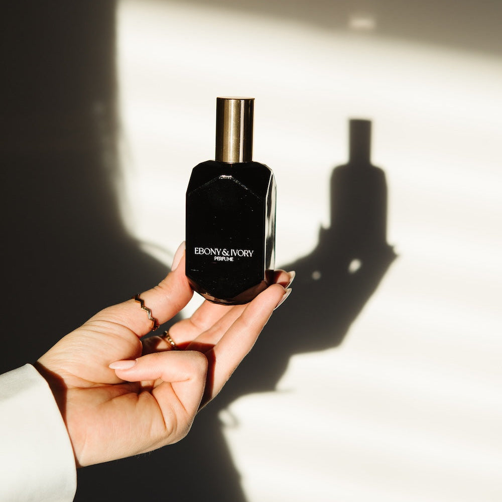 Memoir Collection perfume Debut by Ebony and Ivory. woman's hand holding black perfume bottle with gold lid casting a shadow.