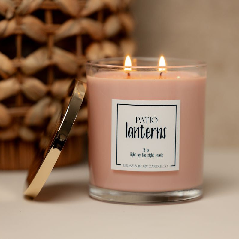A clear jar 16oz candle with a gold lid labelled Patio Lanterns, sitting on a table with a wicker basket. Made in Lethbridge, alberta, canada
