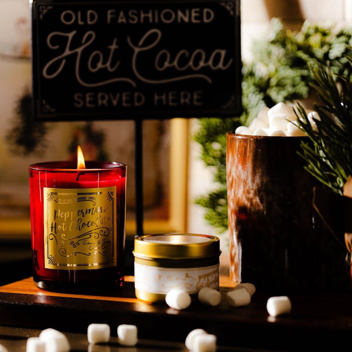 Hot cocoa-themed setup with candles, marshmallows, and a sign in a cozy setting. Red peppermint hot chocolate soy candle for Christmas in a red jar with gold label.