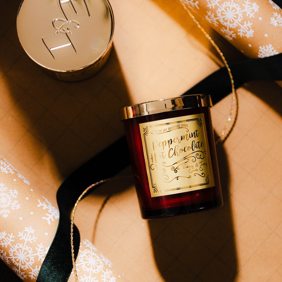 Decorative candle with gold lid and label on a textured surface with gift wrap and ribbon. Red peppermint hot chocolate soy candle for Christmas in a red jar with gold label.
