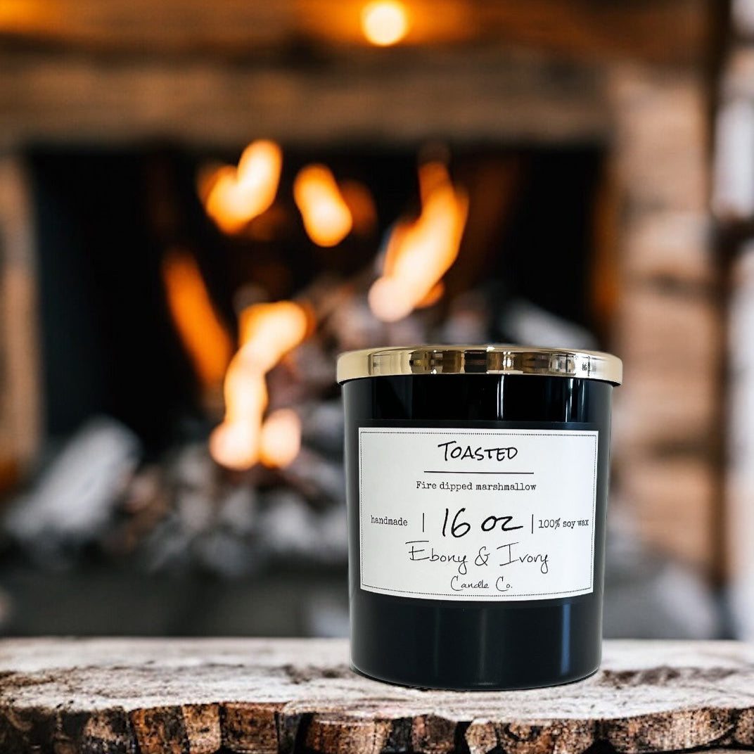 Black candle in a glass container with a label on a wooden surface in front of a fireplace.