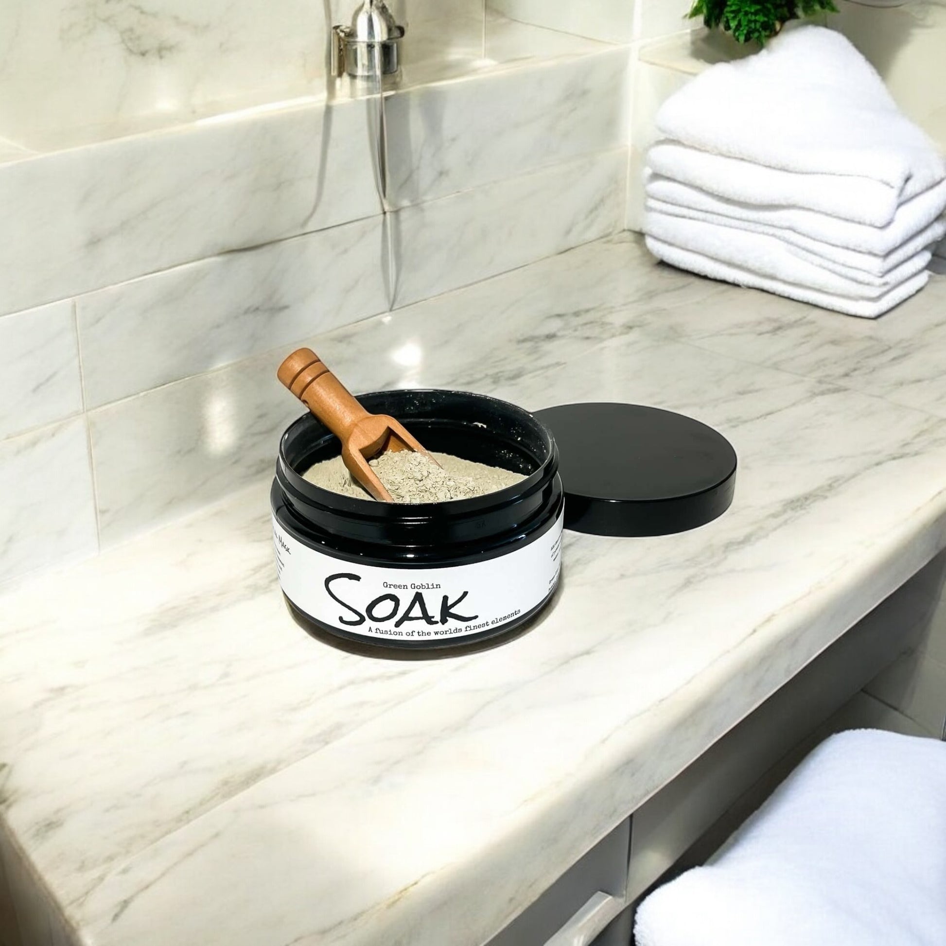 A 4oz round black jar with a white label named Green Goblin Clay Soak, with a wooden spoon, on a white counter top, made by Ebony & Ivory Candle Co in Lethbridge, Alberta, Canada