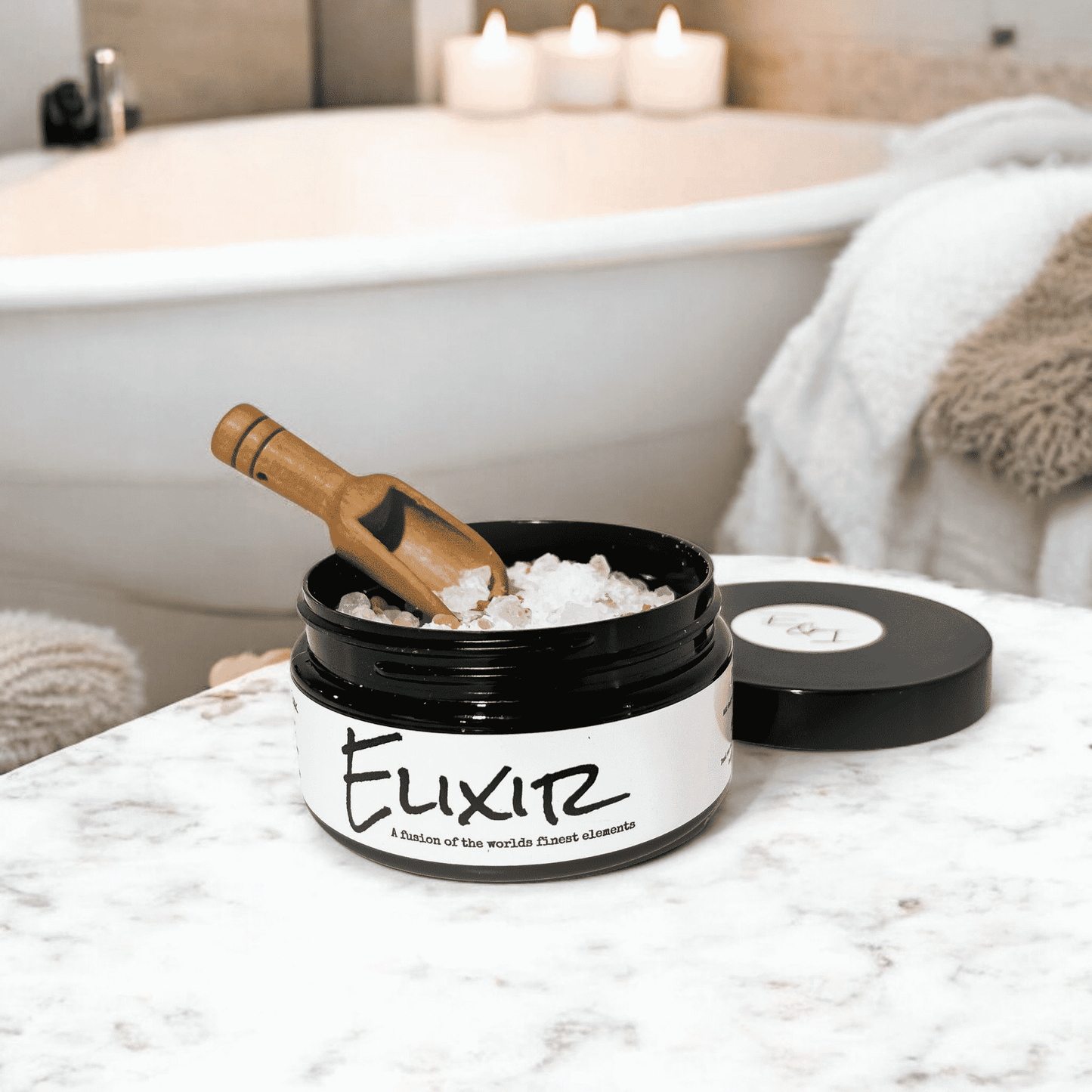 A round Black container with a white label that reads "elixir" and is filled with pink Himalayan salt, dead sea salt, epsom salt, and peppermint essential oils. A wooden spoon is scooping some of the bath soak. Sitting on a white marble counter top in a bathroom. Made by ebony & Ivory candle co in Lethbridge, Alberta, Canada