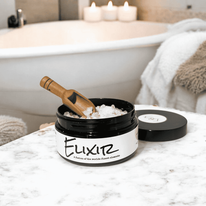 A round Black container with a white label that reads "elixir" and is filled with pink Himalayan salt, dead sea salt, epsom salt, and peppermint essential oils. A wooden spoon is scooping some of the bath soak. Sitting on a white marble counter top in a bathroom. Made by ebony & Ivory candle co in Lethbridge, Alberta, Canada