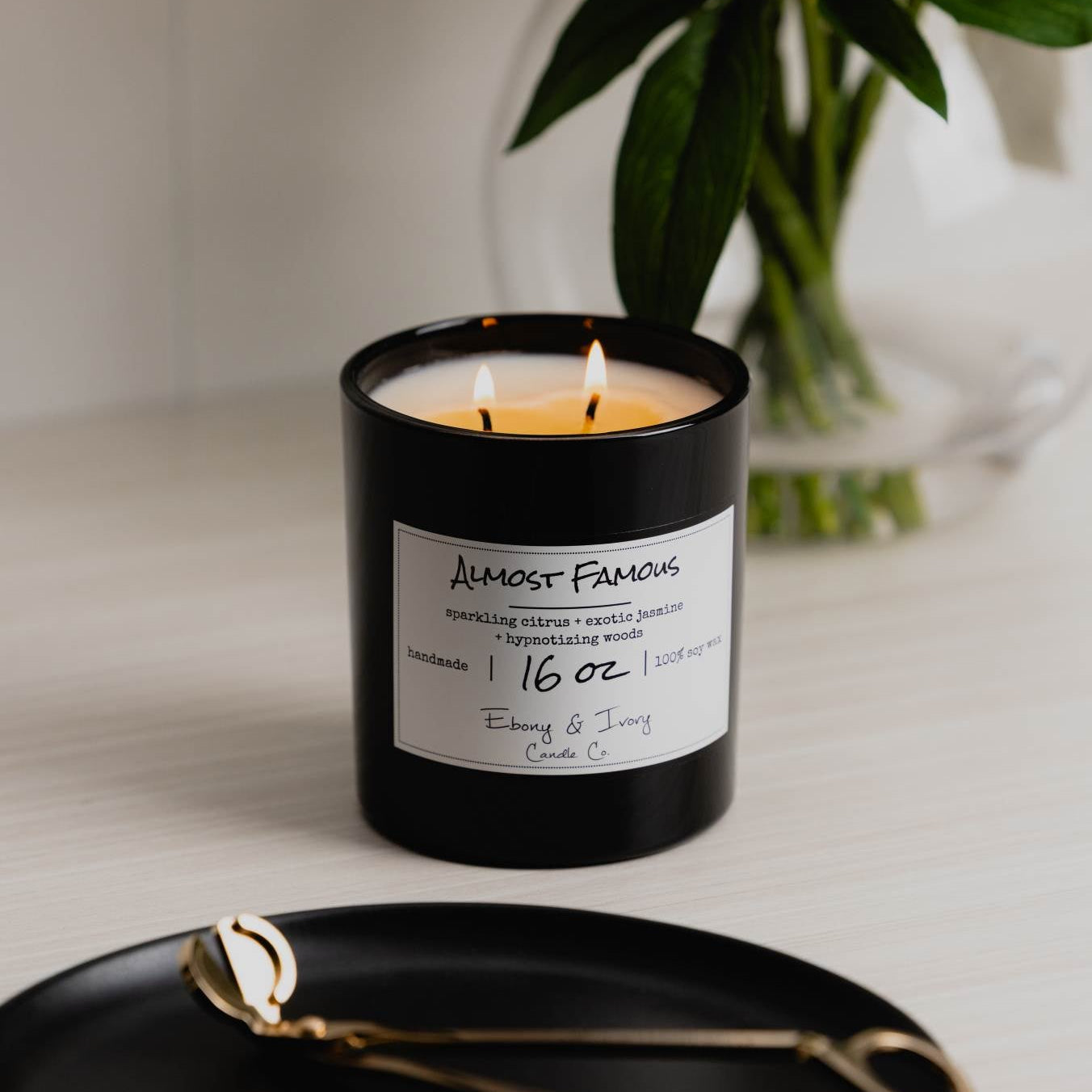 A black 16oz jar soy candle with a white label that reads "Almost Famous" sitting on a clean white table, with a black tray with gold trimmers and a clear vase of flowers in the background, made by Ebony & Ivory Candle Co in Lethbridge, Alberta, Canada