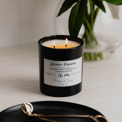A black 16oz jar soy candle with a white label that reads "Almost Famous" sitting on a clean white table, with a black tray with gold trimmers and a clear vase of flowers in the background, made by Ebony & Ivory Candle Co in Lethbridge, Alberta, Canada