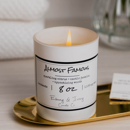 A white 8oz jar soy candle with a white label that reads "Almost Famous" sitting on a gold tray, on a clean white table with fluffy white towels and a clear vase of flowers in the background, made by Ebony & Ivory Candle Co in Lethbridge, Alberta, Canada
