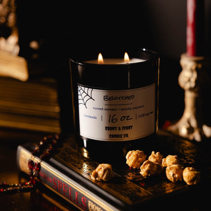 Black 16oz soy candle with a label on a dark surface with books and caramel popcorn, named "bewitched"