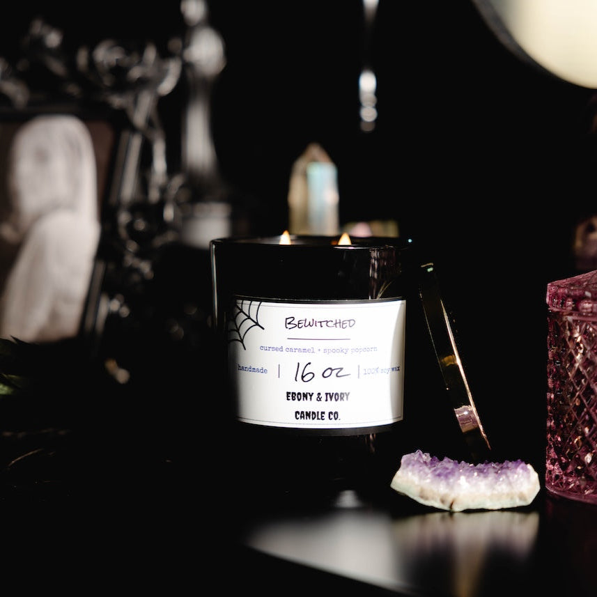 Black 16oz soy candle with a white label on a dark background with decorative items. Named "Bewitched" halloween candle.