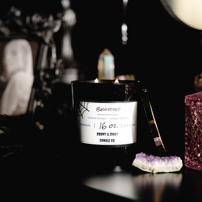 Black 16oz soy candle with a white label on a dark background with decorative items. Named "Bewitched" halloween candle.