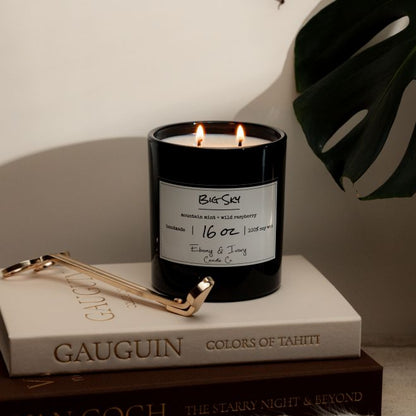 A black 16oz soy candle labelled Big Sky sitting on a stack of books with a pair of gold wick trimmers. Made by Ebony & Ivory Candle Co in Lethbridge Alberta Canada