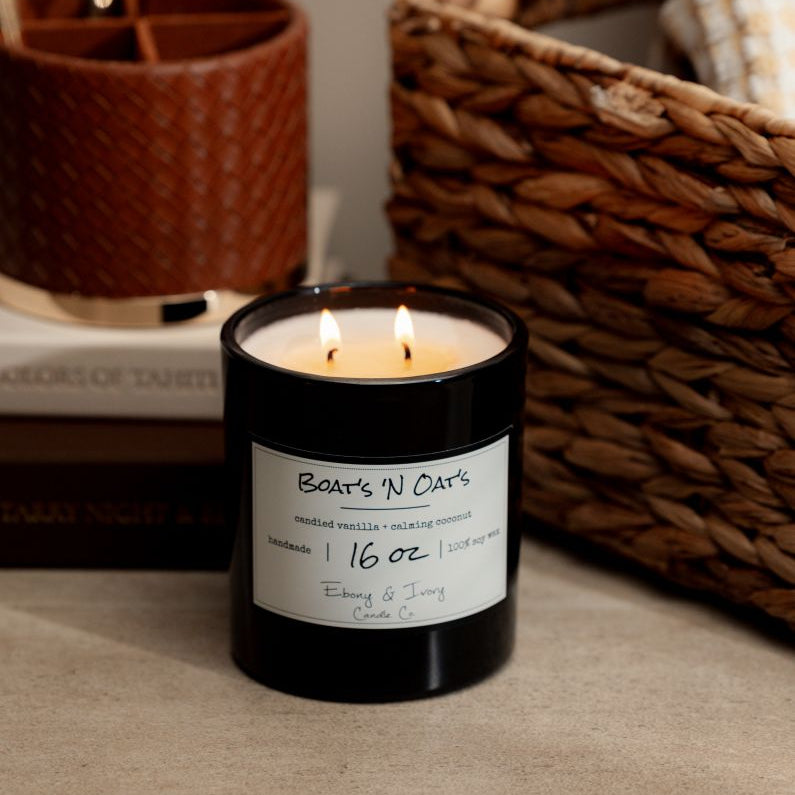 A black 16oz soy candle labelled Boats N Oats, sitting on a table next to artbooks, a pen holder, and a wicker basket. Made by Ebony & Ivory Candle Co in Lethbridge Alberta Canada