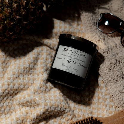 A black 16oz soy candle with a gold lid, labelled Boats N Oats, laying on a beach blanket with sunglasses and a pineapple. Made by Ebony & Ivory Candle Co in Lethbridge Alberta Canada