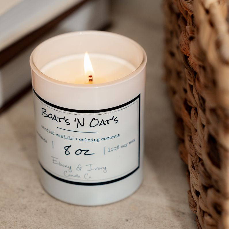 A white 8oz soy candle labelled Boats N Oats, sitting on a table next to books and a wicker basket. Made by Ebony & Ivory Candle Co in Lethbridge Alberta Canada
