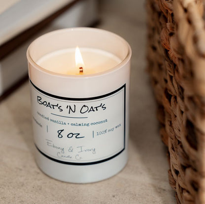 A white 8oz soy candle labelled Boats N Oats, sitting on a table next to books and a wicker basket. Made by Ebony & Ivory Candle Co in Lethbridge Alberta Canada