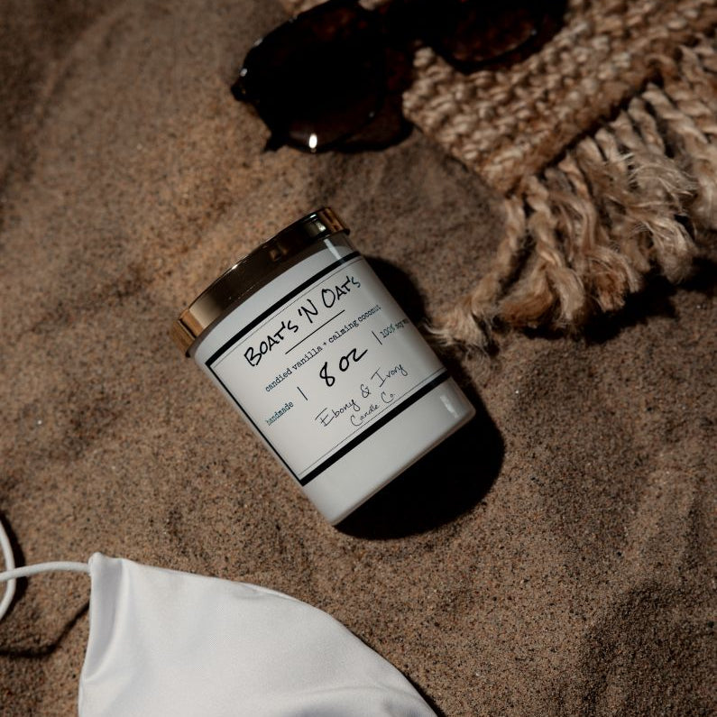 A white 8oz soy candle with a gold lid, labelled Boats N Oats, laying on a beach next to sun glasses, a white bikini top, and a beach blanket. Made by Ebony & Ivory Candle Co in Lethbridge Alberta Canada