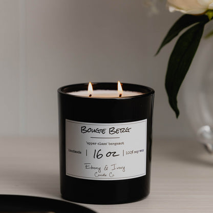 A black 16oz jar soy candle with a gold lid and a white label named Bougie Berg, on a white table with a black tray and a vase of white flowers in the background, made by Ebony & Ivory Candle Co in Lethbridge, Alberta, Canada