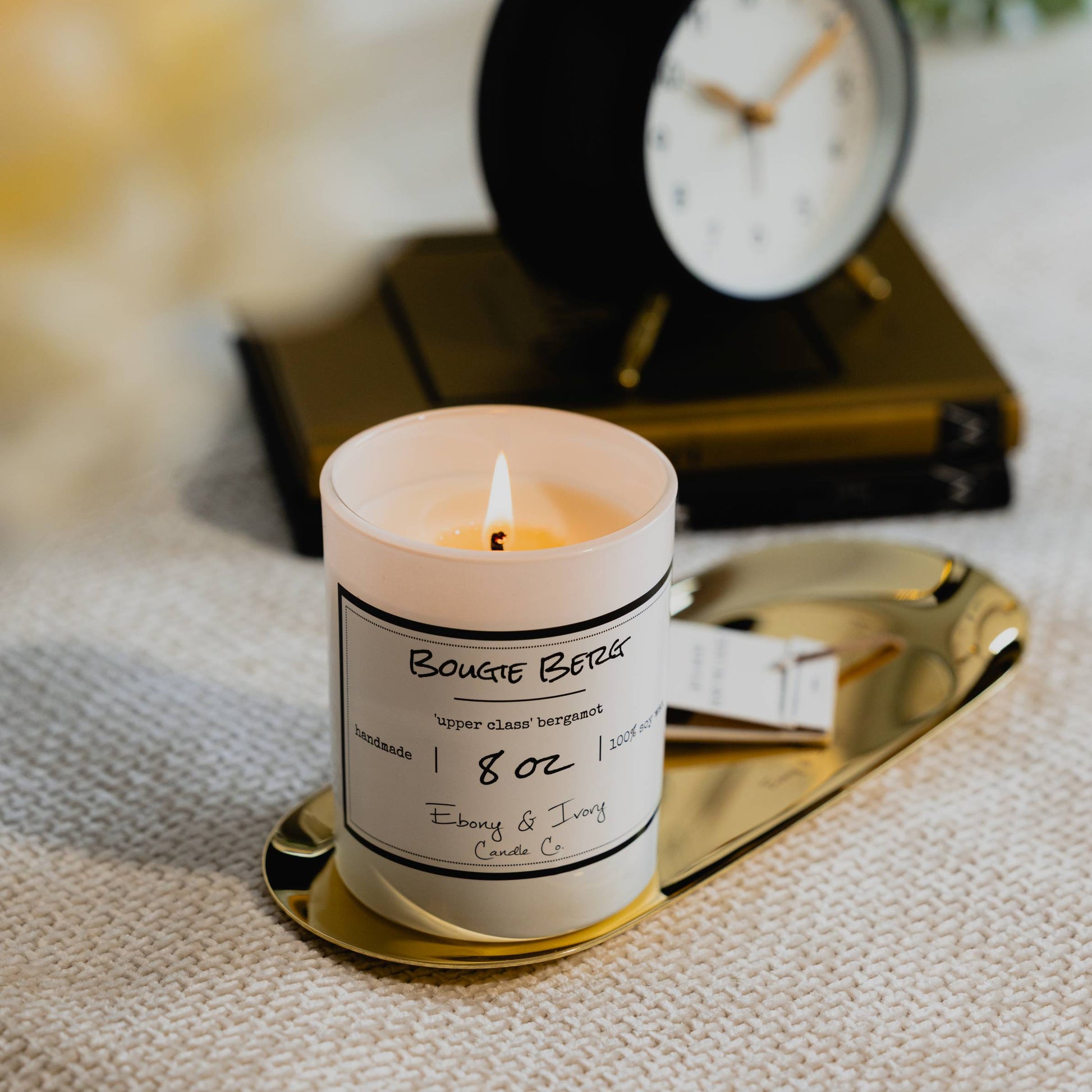 A white 8oz jar soy candle with a white label named Bougie Berg, on a gold tray with a stack of books and a black alarm clock in the background, and white flowers in the foreground, made by Ebony & Ivory Candle Co in Lethbridge, Alberta, Canada