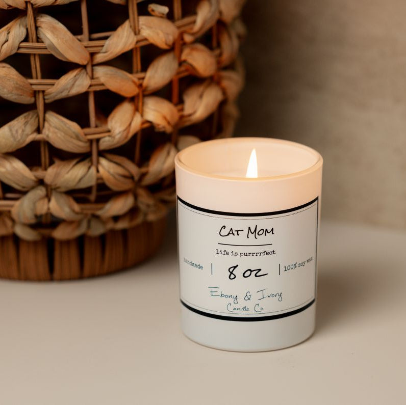 A white 8oz soy candle labelled Cat Mom sitting on a table with a wicker basket behind it. Made in Lethbridge Alberta Canada