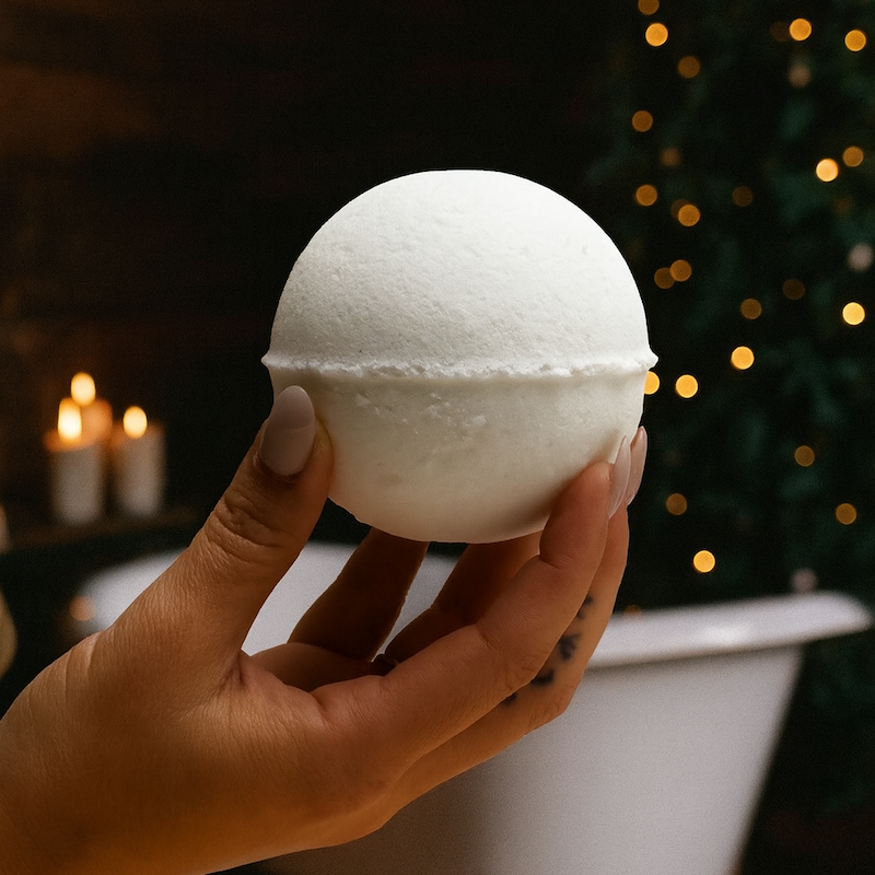 A christmas bathbomb being held up by someone infront of a bathroom with christmas lights, candles, and greenery in the background.