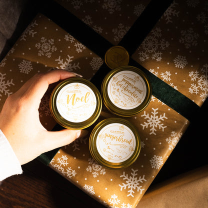 Three small gold 4oz Christmas candles in gold tin with white and gold labels. Candles under the Christmas tree ontop of wrapped presents