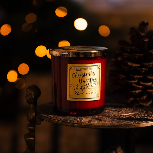 Red 16oz soy christmas candle with a gold lid and gold label named 'Christmas Vacation' on a decorative stand with blurred lights and pine cones in the background. Perfect Christmas candle. 