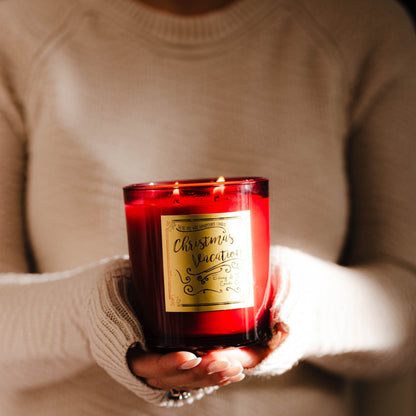 Person holding a red 16oz soy christmas candle with a gold label named "christmas vacation", wearing a beige sweater. Eggnog scented candle called Christmas Vacation.