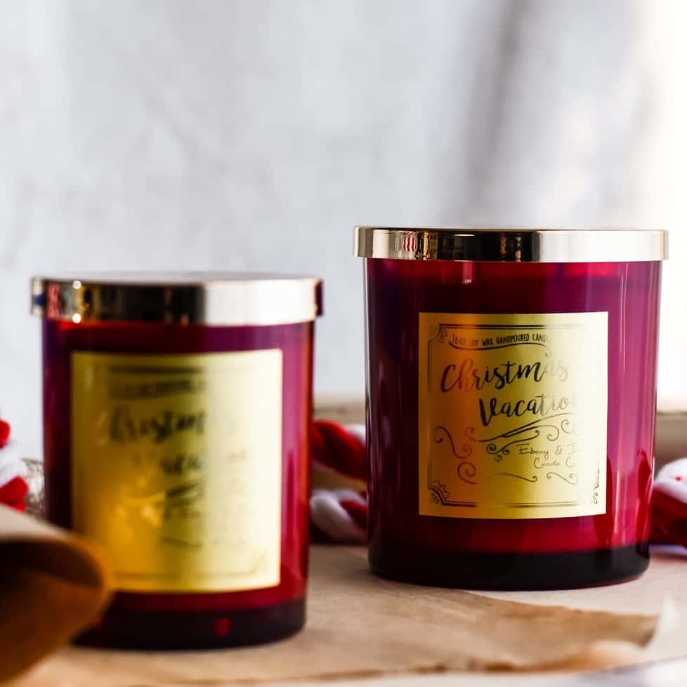 red 8oz and red 16oz soy christmas candles with gold lids and gold lbels named "christmas vacation" sitting on christmas wrapping paper