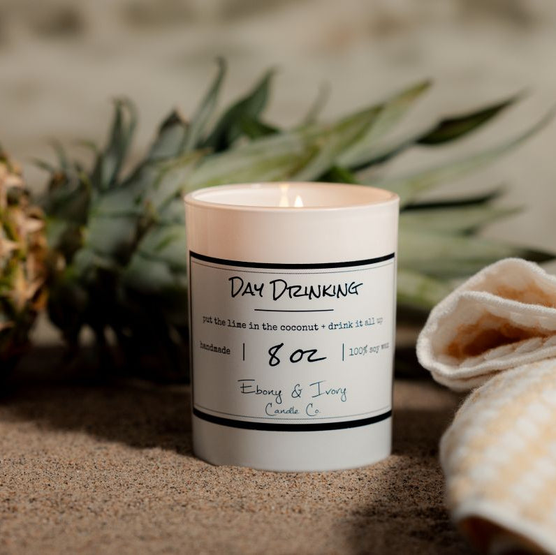 A white 8oz soy candle labelled Day Drinking sitting on the beach with a pineapple and a towel. Made in Lethbridge Alberta Canada