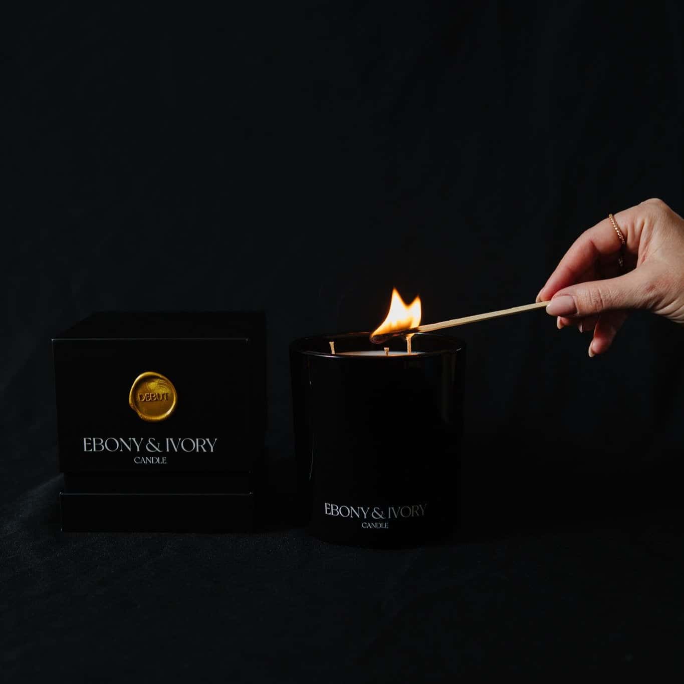 A black 18oz jar coconut soy candle with a white Ebony & Ivory on the front next to a black candle box with a gold label named Debut, on a black background, being lit, made by Ebony & Ivory Candle Co in Lethbridge, Alberta, Canada