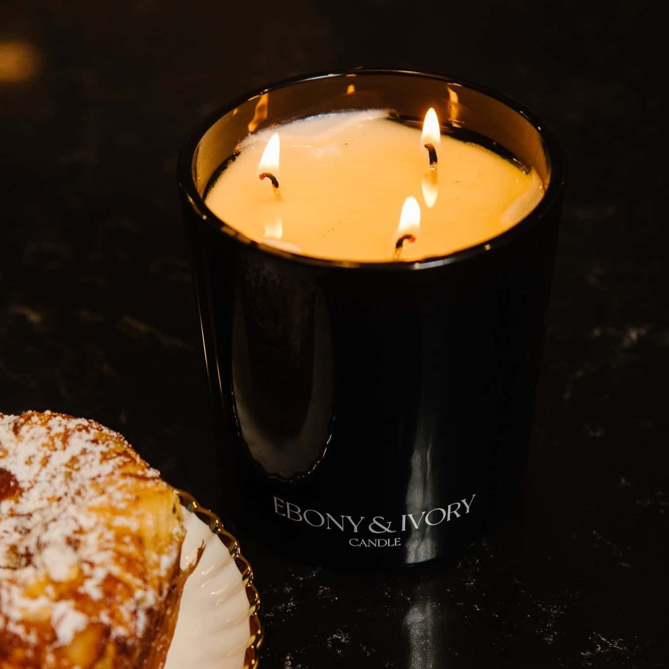 A top down view of a black 18oz jar coconut soy candle with a white Ebony & Ivory on the front of it, next to a plate with a croissant, on a black background, made by Ebony & Ivory Candle Co in Lethbridge, Alberta, Canada