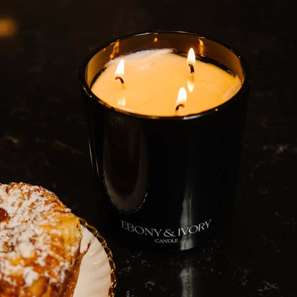 A top down view of a black 18oz jar coconut soy candle with a white Ebony & Ivory on the front of it, next to a plate with a croissant, on a black background, made by Ebony & Ivory Candle Co in Lethbridge, Alberta, Canada