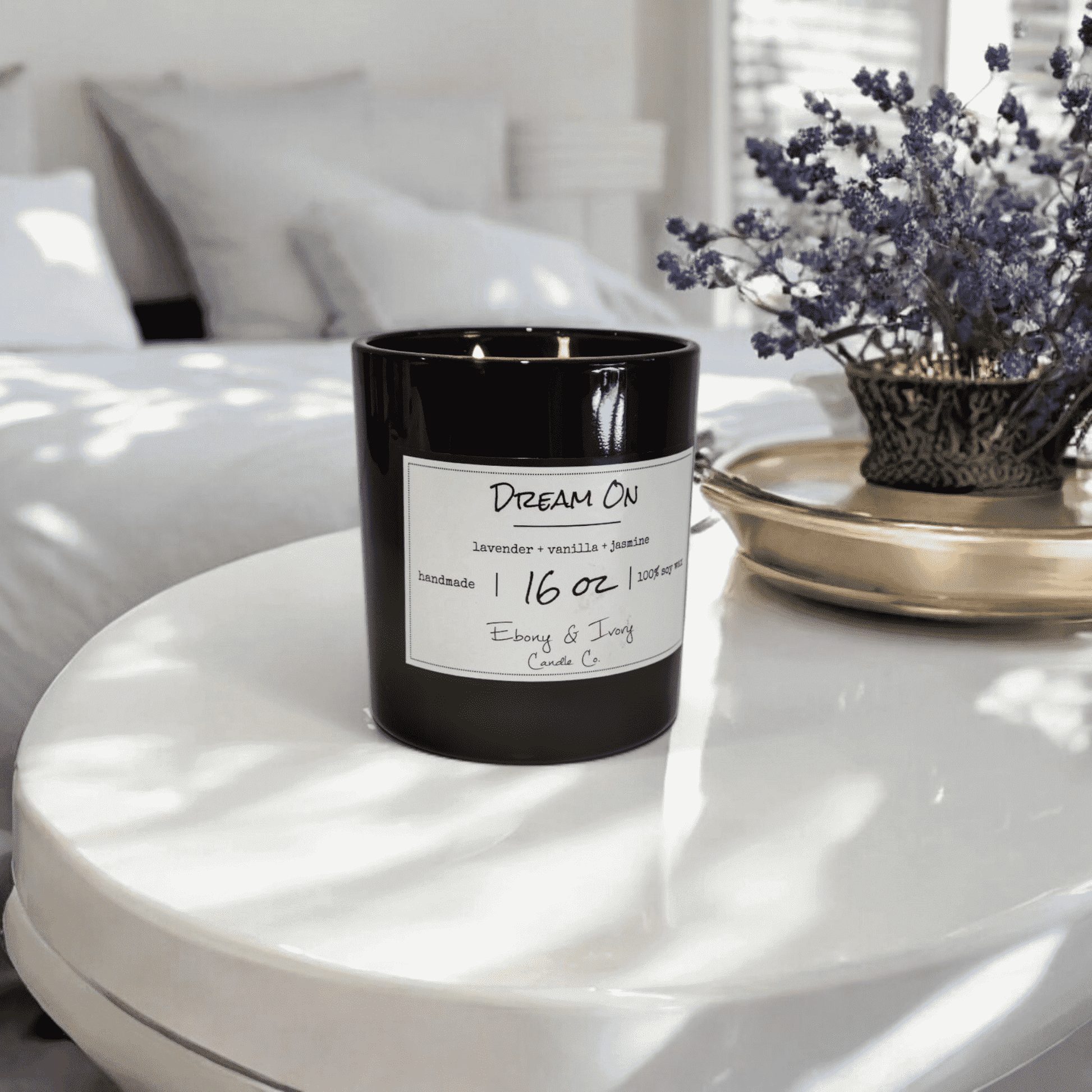 A black 16oz jar soy candle with a gold lid and a white label named Dream On, on a white table with a white bed and a pot of lavender in the background, made by Ebony & Ivory Candle Co in Lethbridge, Alberta, Canada