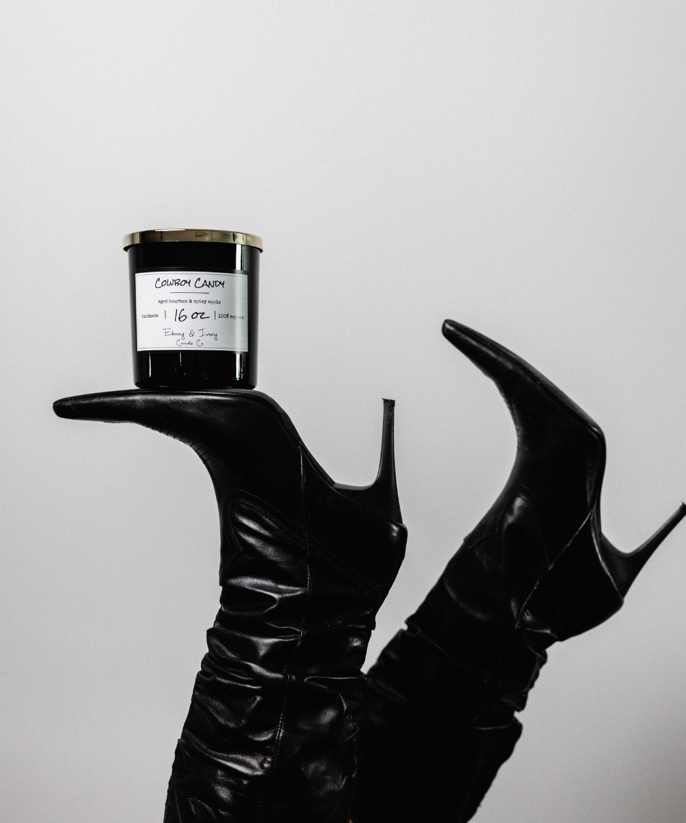 Black leather high-heeled shoes holding a black candle with a white label against a light gray background