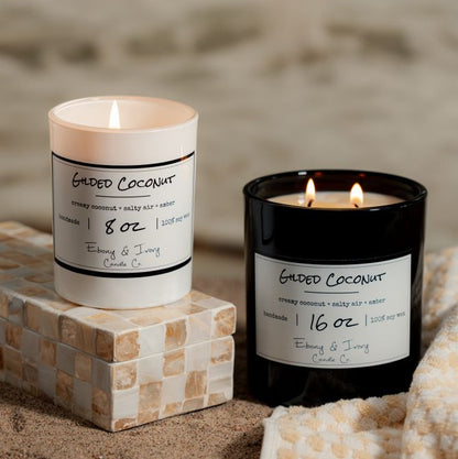 A white 8oz and black 16oz soy candles labelled 'Gilded Coconut' sitting on the beach. Made in Lethbridge Alberta Canada