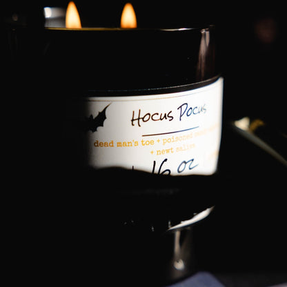 Black 16oz soy candle with a white label named "hocus pocus" on a dark spooky background