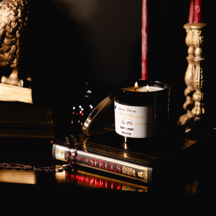 Black 16oz Soy candle with a label on books and a statue in a dark romantic setting. named "hocus pocus"