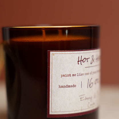 A close up shot of the side of a 16oz soy candle in an amber jar with a lable that reads "Hot & Heavy"