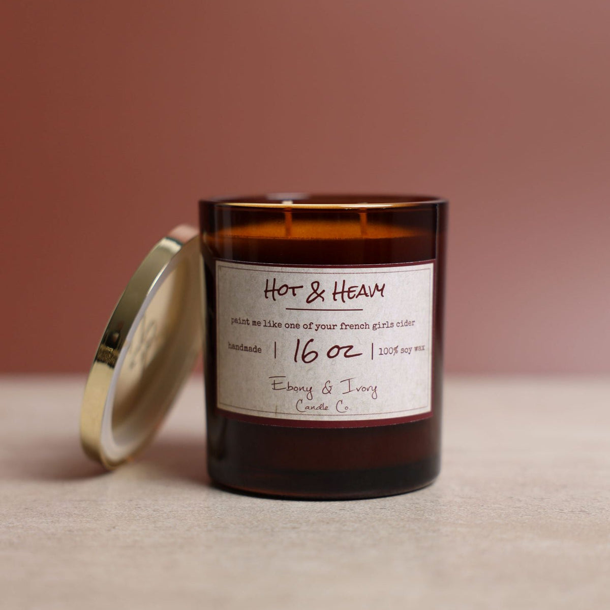 An amber 16oz jar soy candle with a gold lid and a white label named "Hot & Heavy" sitting on a table.